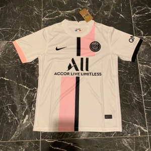 PSG vit och rosa matchtröja Nike - Paris Saint-Germain fotbollströja i vitt med en rosa och svart vertikal rand på framsidan. Tröjan har korta ärmar, svart Nike-logga och PSG-emblem på bröstet. Materialet är lätt och andas, perfekt för träning eller match.