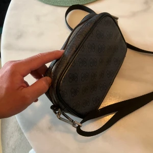 Svart axelväska från Guess med mönster - Säljer en svart axelväska från Guess med justerbar rem och silverfärgade detaljer. Väskan har Guess-logga framtill och ett diskret mönster över hela ytan. Tillverkad i syntetmaterial med dragkedja och praktisk form, perfekt för att bära det viktigaste.
