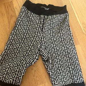 Svartvita mönstrade monogram bikershorts från Balmain - Snygga shorts från Balmain med svartvitt grafiskt mönster och bred resår i midjan. De har en loose passform och är perfekta för dig som vill sticka ut med en lyxig vibe. Tillverkade i ett mjukt material som känns skönt mot huden. Saknar Balmaintagg och säljs därav till priset, samt att lappen där i är lite lös. Annars i perfekt skick.