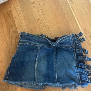 Mini jeanskjol med spännen från Dollhouse - Cool blå jeanskjol från Dollhouse med rå kant nertill och flera snygga spännen i sidan. Kjolen har bakfickor med broderade detaljer och dragkedja bak. Perfekt för dig som gillar en edgy och unik stil. Står storlek 7 inuti men skulle säga att det är en storlek 34/36. 
