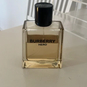 Burberry Hero Eau de Toilette 100ml - Burberry Hero Eau de Toilette i en stilren, fyrkantig glasflaska med svart plastkork. Flaskan rymmer 100 ml och har en ljusgul vätska. Märkets logga syns tydligt på framsidan. Perfekt för dig som gillar klassiska och moderna dofter.