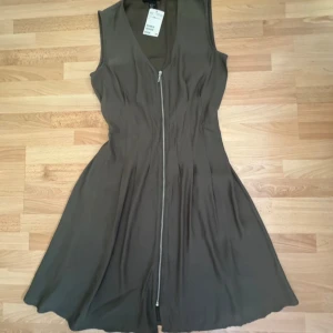 Ny. Olivgrön ärmlös klänning med dragkedja - Olivgrön ärmlös klänning från Vero Moda med v-ringning och hel dragkedja framtill. Klänningen har en lätt utställd kjol och markerad midja med resår baktill. Perfekt för dig som gillar enkel och stilren design.