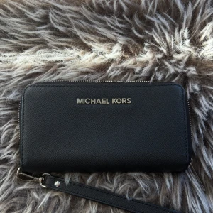 Svart plånbok från Michael Kors - Snygg svart plånbok från Michael Kors med silverfärgad logga framtill. Plånboken har avtagbar handledsrem, dragkedja runtom och flera fack för kort och annat. Tillverkad i strukturerat skinn som ger en lyxig känsla och stilren look.