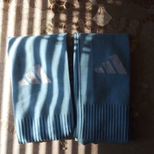 Blåvita randiga Adidas strumpor - Säljer ett par blåa knästrumpor från Adidas med vita ränder och klassisk logga. Strumporna är ribbstickade nertill och har en sportig vibe, perfekta till träning eller fotboll. Materialet känns som bomullsmix och de sitter skönt på benet.