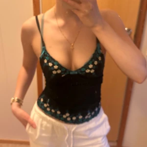 Svart blommig linnetopp med spets - Supersöt svart linnetopp med tunna axelband och broderade blommor i turkos och vitt. Toppen har spetsdetaljer längs kanterna och en liten rosett framtill. Sitter tight och är croppad, perfekt till jeans eller kjol för en trendig look.