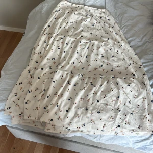 Blommig långkjol från lager 157 - Supersöt långkjol i vitt med små blommor i blått, rosa och grönt från lager 157. Kjolen har resår i midjan och volangiga lager som ger en flowy känsla. Perfekt för dig som gillar romantisk och somrig stil. Frakt ingår i priset 