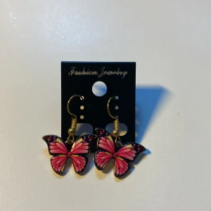 Handgjorda, unika Rosa fjärilsörhängen med gulddetaljer *Endast 1 Kvar* - Supersöta örhängen med fjärilar i rosa och svarta toner, hängande på guldfärgade krokar. Fjärilarna har fina detaljer och en lekfull design som verkligen sticker ut. Perfekta accessoarer för dig som vill ha något unikt och färgglatt.