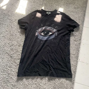 Svart Kenzo t-shirt med ögontryck - Svart t-shirt från Kenzo med ikoniskt ögonmotiv och texten 'Kenzo Paris' på bröstet. Klassisk passform och rund halsringning. Perfekt för dig som vill sticka ut med en cool och unik design. Stlk M men väldigt liten, mer som en XS. Inga tecken på användning.