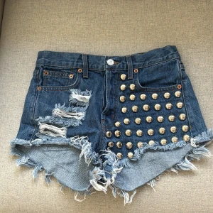 Blå jeansshorts med nitar och slitningar - Snygga blå jeansshorts med råa, fransiga kanter och slitningar framtill och baktill. Ena framsidan är täckt med stora silvriga nitar för en edgy look. Klassisk femficksmodell med bälteshällor och knappstängning. Perfekta för dig som vill sticka ut i sommar!