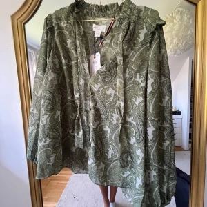 Grön mönstrad blus Holly & White - Snygg blus från Holly & White i grönt paisleymönster. Luftig modell med långa ärmar, volangkrage och knappar framtill samt vid ärmslut. 💕 lapp kvar