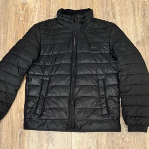 Dunjacka   - Svart pufferjacka från BOSS med horisontella sömmar, dragkedja framtill och två fickor med knappdetalj. Jackan har en stilren look och är tillverkad i syntetmaterial, perfekt för kyliga dagar. Klassisk passform och diskret BOSS-logga på bröstet.                                                                       Priset kan disckuteras