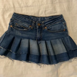Jeanskjol med volang från Noisy May - Supersöt kort jeanskjol från Noisy May med volangkant och råa kanter. Klassisk blå denim med fickor fram och bak, bälteshällor och knäppning framtill. Perfekt för dig som vill ha en edgy och trendig look.