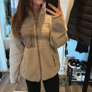 Beige teddyjacka med dragkedja - Mysig beige teddyjacka med hög krage och tre fickor med dragkedja, varav en på bröstet i kontrasterande tyg. Jackan har en relaxed passform. Från lager157 för 2 år sen men aldrig använd🌸
