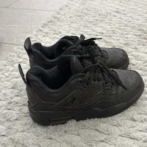 Svarta Nike Air Jordan 4 sneakers - Säljer ett par helsvarta Nike Air Jordan 4 sneakers med klassisk mesh och detaljer i skinn. Skorna har snörning, perforerad tå och den ikoniska Jumpman-loggan på hälen. Perfekta för dig som gillar streetstyle och vill ha en clean look.