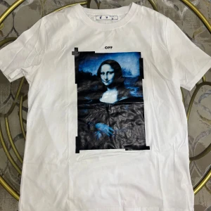 Vit Off-White t-shirt med tryck - Vit t-shirt från Off-White med stort grafiskt tryck framtill i blått och svart, samt de ikoniska svarta pilarna på ryggen. Klassisk passform och rund halsringning. Perfekt statement-plagg för dig som gillar streetwear och unika prints.