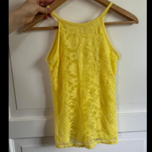 Gul spetsig linnetopp från D-XEL - Superfin gul linnetopp från D-XEL med smala axelband och helspets över hela toppen. Toppen har en liten knapp i nacken och är ärmlös med en rak passform. Perfekt för dig som vill sticka ut med en färgglad och somrig look!