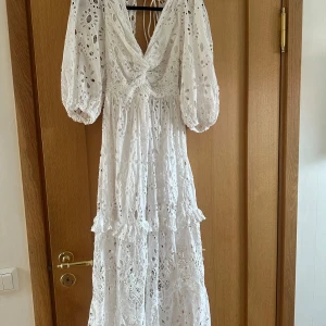 Vit långklänning med broderade detaljer - Superfin vit långklänning med broderade blommor och hålmönster. Klänningen har v-ringning, puffiga trekvartsärmar och volangdetaljer. Öppen i ryggen med band! Använd fåtal gånger
