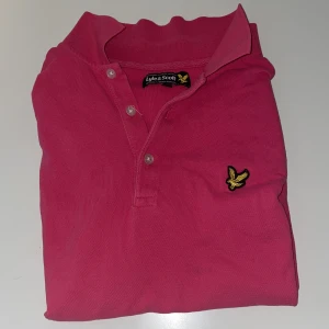 Rosa pikétröja från Lyle & Scott - Snygg rosa pikétröja från Lyle & Scott med klassisk krage, knappar och det ikoniska gula örnloggan på bröstet. Tröjan har korta ärmar, perfekt för en chill och stilren look.