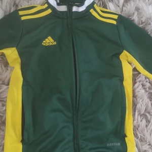 Adidas grön och gul tröja  - Sportig tröja från Adidas i grönt med gula detaljer och klassiska ränder på axlarna. Jackan har hel dragkedja, lång ärm och är tillverkad i lätt polyester. Perfekt för träning eller när du vill ha en cool streetlook.
