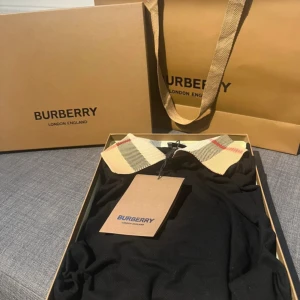 Svart Burberry pikétröja med rutig krage - Snygg svart pikétröja från Burberry med klassisk rutig krage i beige, svart och rött. Tröjan har korta ärmar och är tillverkad i bomull. Perfekt för dig som vill ha en stilren och lyxig look med en touch av ikoniskt mönster.