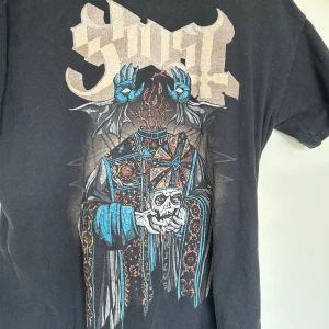 Svart Ghost band t-shirt - Svart t-shirt från Gildan med stort färgstarkt Ghost-bandtryck på framsidan. Klassisk passform med korta ärmar och rund hals. Merch från typ 2020. Helt felfri (men ksk lite katthår på bilderna)
