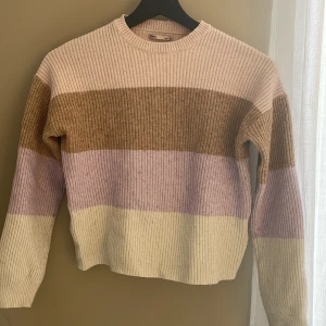Randig stickad tröja från ONLY - Supermysig stickad tröja från ONLY med breda ränder i beige, ljusrosa, lila och brunt. Tröjan har ribbad struktur, rund hals och långa ärmar. Perfekt för dig som gillar en chill och färgglad stil.