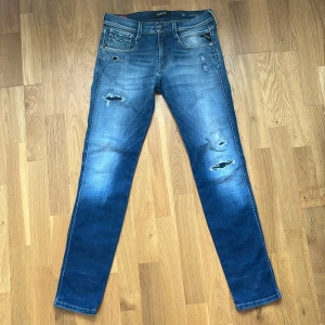 Replay blå slitna jeans W29 L32 - Snygga blå jeans från Replay med slitna detaljer och lätt tvättad look. Modellen har klassisk femficksdesign, normal midja och raka ben. Jeansen är tillverkade i bomull med en skön passform och coola slitningar på framsidan.