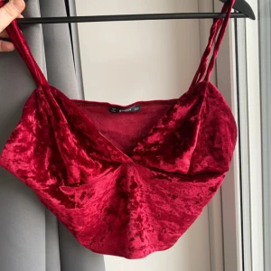 Vinröd sammetstopp från SHEIN - Säljer en vinröd croppad topp från SHEIN i mjuk sammet. Toppen har omlott-skärning framtill, smala axelband och en snygg glansig finish. Perfekt för dig som vill sticka ut med en djup v-ringning och trendig look.