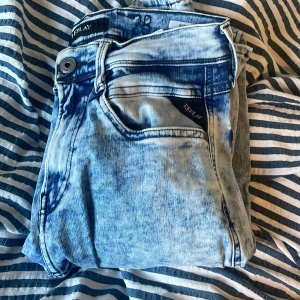 Replay Anbass ljusblå jeans, stl 28 - Säljer ett par Replay Anbass jeans i en snyggt ljusblå tvätt med slitna detaljer. Klassisk femficksmodell med Replay-logga vid fickan och knappstängning. Jeansen har en modern passform och är perfekta för dig som gillar en fräsch och trendig look.