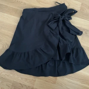 Svart omlottkjol med volang och rosett - Supersöt svart omlottkjol med volangkant och stor rosett i midjan. Tunnt material och perfekt att bära under varmare dagar. Storlek xs men passar även S då den har stretch i midjan. 