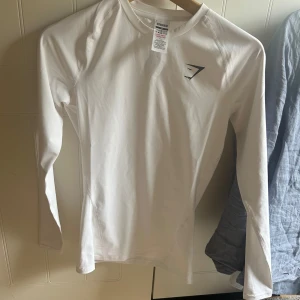 Vit långärmad träningströja Gymshark - Säljer en vit långärmad träningströja från Gymshark i storlek XS. Tröjan har en tight passform och är tillverkad i mjuk polyester, perfekt för gymmet eller löprundan. Diskret svart Gymshark-logga på bröstet och stilrena sömmar för sportig look. Gymshark element baselayer long sleeve t shirt compression fit, säljes då jag fick fel storlek och ej kan returnera. Helt ny inte ens testad