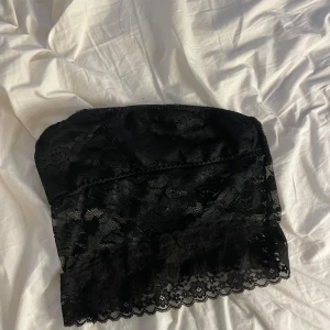 Svart spets bandeau tubtopp - Säljer en svart bandeau tubtopp i spets med blommigt mönster. Toppen är axelbandslös och har en fin, vågig spetskant nedtill. Perfekt för att ge en extra touch till din outfit. Mjuk och stretchig för skön passform.