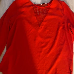 Röd blus med spetsdetalj - Snygg röd blus med lång ärm och v-ringning framtill som har en transparent spetsdetalj. Perfekt för dig som vill ha något stilrent men ändå med en twist. Blusen har en rak passform och är tillverkad i ett mjukt tyg som känns skönt mot huden.