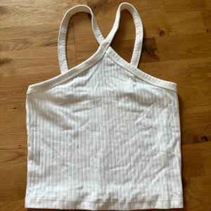 Vit ribbad linnetopp med korsade band - Snygg vit ribbad linnetopp med smala axelband som korsas i ryggen. Toppen är croppad och har en enkel, stilren look som passar perfekt till jeans eller kjol. Tillverkad i mjuk bomull för en skön känsla hela dagen.