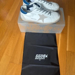 Golden Goose  - Golden Goose sneakers i vitt skinn med grå mockadetaljer och blå häl. Klassisk stjärna på sidan, grå snörning och platt sula. Snygg och avslappnad modell som passar perfekt till jeans eller shorts. Kommer med originalkartong och dustbag.🙌🏻 STORLEK 40 sitter mer som 41 skulle jag säga.