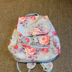Blommig ryggsäck från Cath Kidston - Superfin ryggsäck från Cath Kidston i ljusblått med stora blommor i rosa, rött och lila. Väskan har flera fack, justerbara axelband och ett spänne framtill. Perfekt för skolan eller utflykter, och den blommiga designen gör den extra söt.
