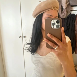 Beige basker från Wera i ull - Stilren beige basker från Wera i mjuk ull. Klassisk rund form med liten topp och diskret gulddetalj framtill. Perfekt accessoar för att lyfta din look med fransk vibe. 