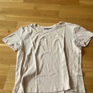 Beige basic t-shirt från Zara - Enkel och stilren beige t-shirt från Zara i mjuk bomull. Klassisk rund halsringning och korta ärmar. Perfekt att matcha med jeans eller shorts för en clean look.
