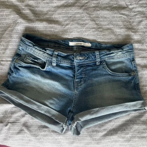 Blå jeansshorts från Sinsay - Klassiska blå jeansshorts från Sinsay med upprullade benslut och femficksmodell. Shortsen har normal passform och bälteshällor, perfekt till sommaren. Snyggt slitna detaljer och dragkedja framtill ger en avslappnad vibe.