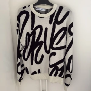 Vit och svart tröja från Bershka - Snygg vit tröja från Bershka med stora svarta grafiska bokstäver över hela plagget. Rund halsringning och ribbade muddar vid ärmslut och nederkant. Perfekt för dig som gillar statement-plagg och streetstyle.