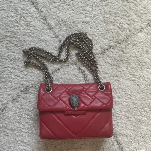 Röd quiltad axelväska Kurt Geiger - Säljer en snygg röd axelväska från Kurt Geiger i quiltat skinn. Väskan har en cool silverfärgad kedjerem och ikoniskt fågelhuvud som spänne. Perfekt storlek för det viktigaste och ger en edgy vibe till din outfit.