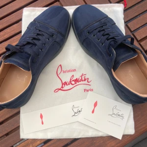 Mörkblå mocka sneakers från Louboutin - Snygga mörkblå sneakers från Christian Louboutin i mjuk mocka med klassisk röd sula. Skorna har rund tå, blåa snören och diskret broderad logga på hälen. Insidan är i ljust skinn för extra komfort. Perfekta för dig som vill ha en lyxig och stilren look. Tar emot byten