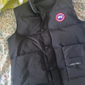 Svart dunväst från Canada Goose - Säljer en svart dunväst från Canada Goose med klassisk logga på bröstet. Västen har stora fickor framtill, dragkedja och tryckknappar. Perfekt för kalla dagar och har en clean, stilren look som funkar till det mesta.