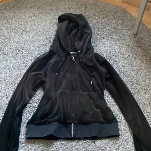 Svart velour hoodie från Juicy Couture - Svart hoodie i mjuk velour från Juicy Couture med dragkedja, huva och snörning. Klassisk logga broderad på bröstet och känguruficka framtill. Perfekt för en chill och trendig look. Storlek xxs