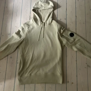 Beige hoodie från C.P. Company - Helt ny! Snygg beige hoodie från C.P. Company med klassisk linsdetalj på ärmen. Tröjan har huva med snörning och är tillverkad i mjuk bomull. Perfekt för dig som gillar stilrena och avslappnade plagg med en twist. Oanvänd, tags finns nånstans, kan letas upp på begäran. 