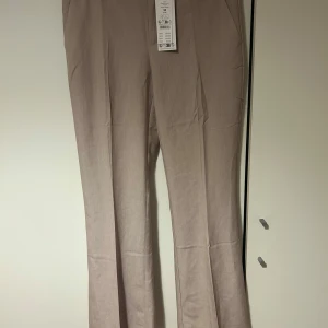 Beige kostym byxor  - Snygga beige melerade byxor med bootcut-snitt och relaxed fit. Byxorna har en klassisk look med hög midja och är tillverkade i en linneblandning som ger en lätt och luftig känsla. Perfekta för dig som gillar stilrena och bekväma byxor.