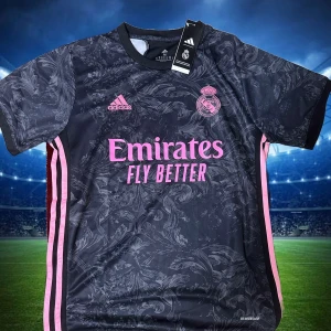 Real Madrid svart bortatröja Adidas - Snygg svart Real Madrid bortatröja från Adidas med rosa detaljer och logga. Tröjan har korta ärmar, subtilt mönster över hela framsidan och rosa ränder längs sidorna. Tillverkad i lätt och ventilerande material, perfekt för fotbollsplanen storlek S,M.