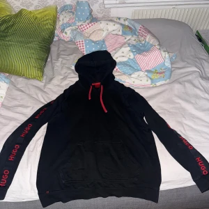 Svart hoodie från Hugo med röda detaljer - Svart hoodie från Hugo med röda snören och röd logga längs båda ärmarna. Klassisk känguruficka framtill och huva. Skön passform och stilren look, perfekt för dig som gillar streetwear och vill sticka ut lite extra.