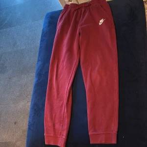 Vinröda mjukisbyxor från Nike XL - Vinröda mjukisbyxor från Nike med resår i midjan och dragsko. Byxorna har en klassisk Nike-logga i vitt på ena benet och är tillverkade i mjukt bomullsmaterial. Perfekta för chill eller träning, med bekväm passform och muddar vid bensluten. Det finns jacka till i annan annons!