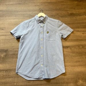 Ljusblå kortärmad skjorta Lyle & Scott - Snygg ljusblå skjorta från Lyle & Scott med korta ärmar och klassisk button down-krage. Skjortan har bröstficka med den ikoniska gula örnloggan och stängs med vita knappar. Perfekt för en clean och stilren look.